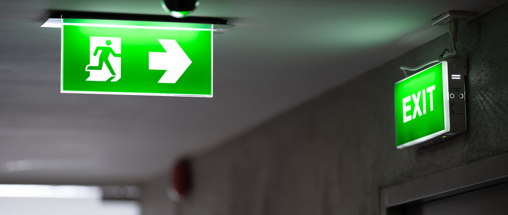 A green fire exit sign is placed on the ceiling along the dimly lit corridor and there is green exit sign on the exit door.